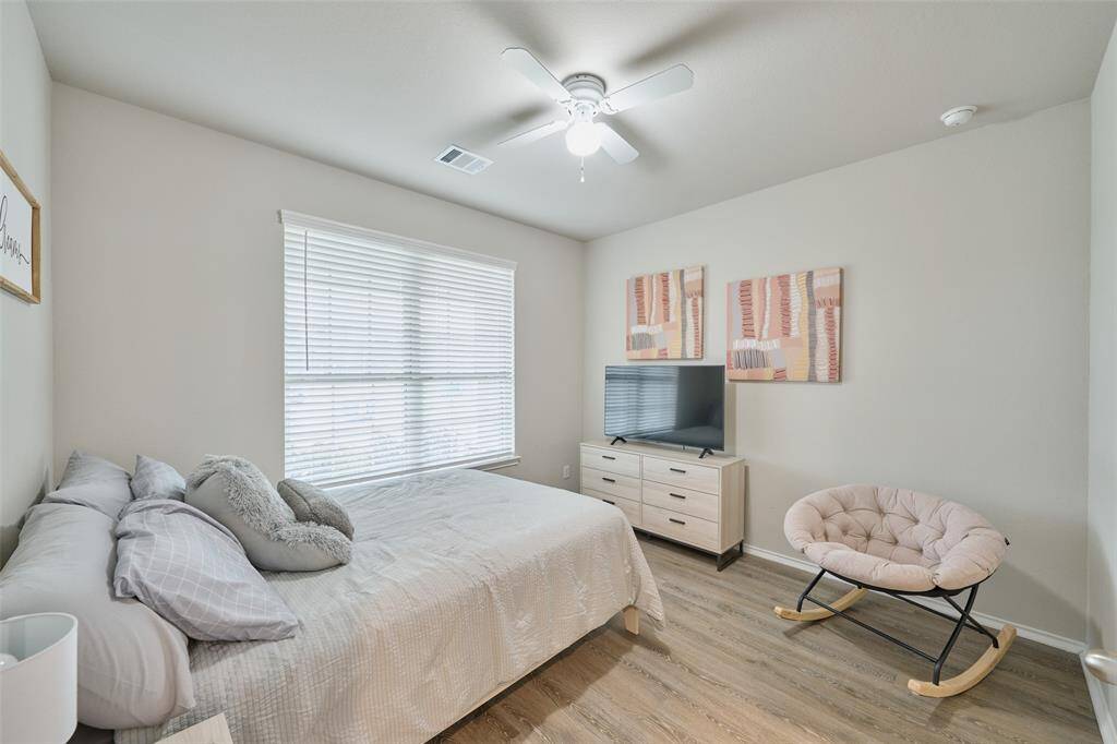 Cozy bedroom with natural light, featuring a bed, modern dresser with a TV, and an accent chair. Light wood flooring and neutral walls create a calming atmosphere.