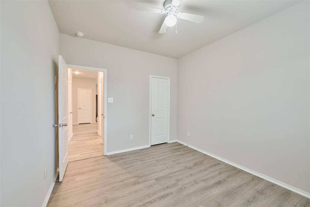 This room features light gray walls, wood-style flooring, and a ceiling fan. It has a door leading to a hallway, offering a clean and bright space, ideal for a bedroom or office.