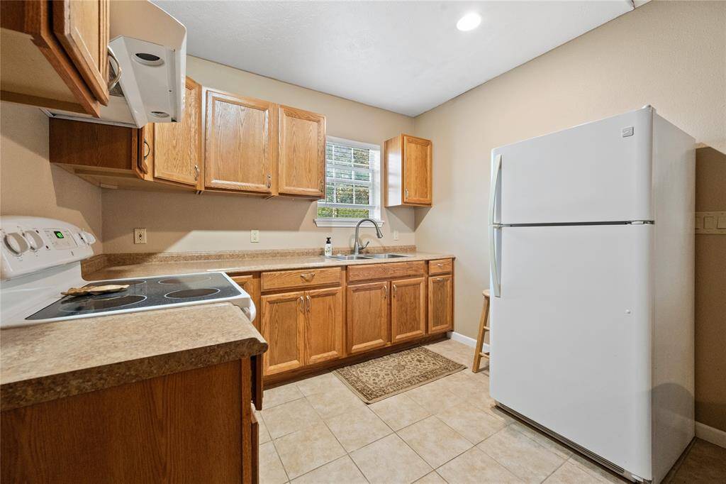 Guest house kitchen with wooden cabinets, a white refrigerator, and an electric stove. Features a window above the sink providing natural light, and neutral tile flooring.