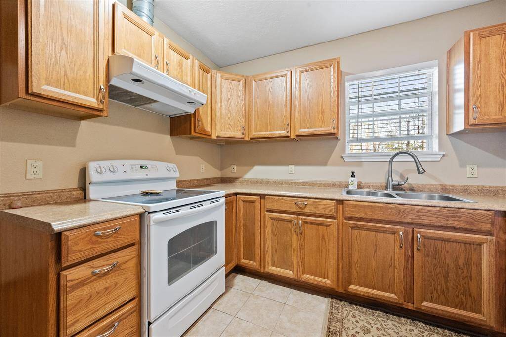 Guest house kitchen with wooden cabinets, a white refrigerator, and an electric stove. Features a window above the sink providing natural light, and neutral tile flooring.