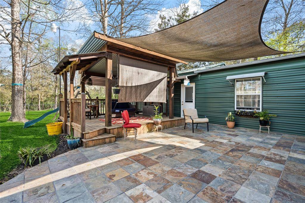 This photo showcases a charming outdoor space with a tiled patio, shaded seating area, and a small deck. The green siding of the house complements the natural surroundings, and there's a slide in the lush backyard, ideal for family activities.