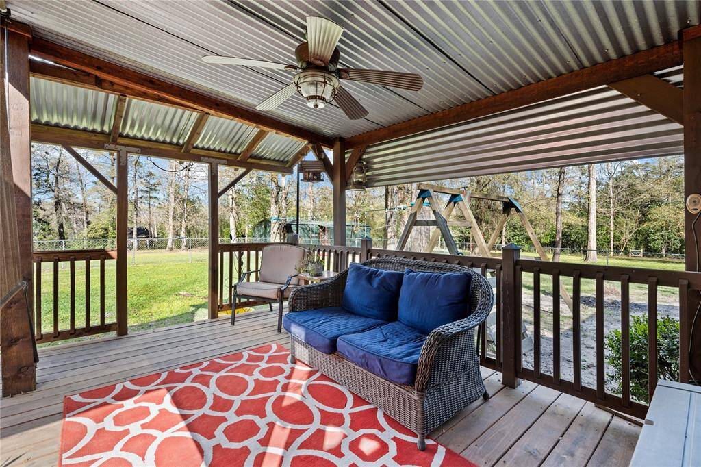 This photo showcases a charming covered pergola with a metal roof and wooden deck. It features comfortable outdoor seating, a ceiling fan, and a vibrant rug, offering a cozy space to enjoy the spacious, tree-lined backyard.