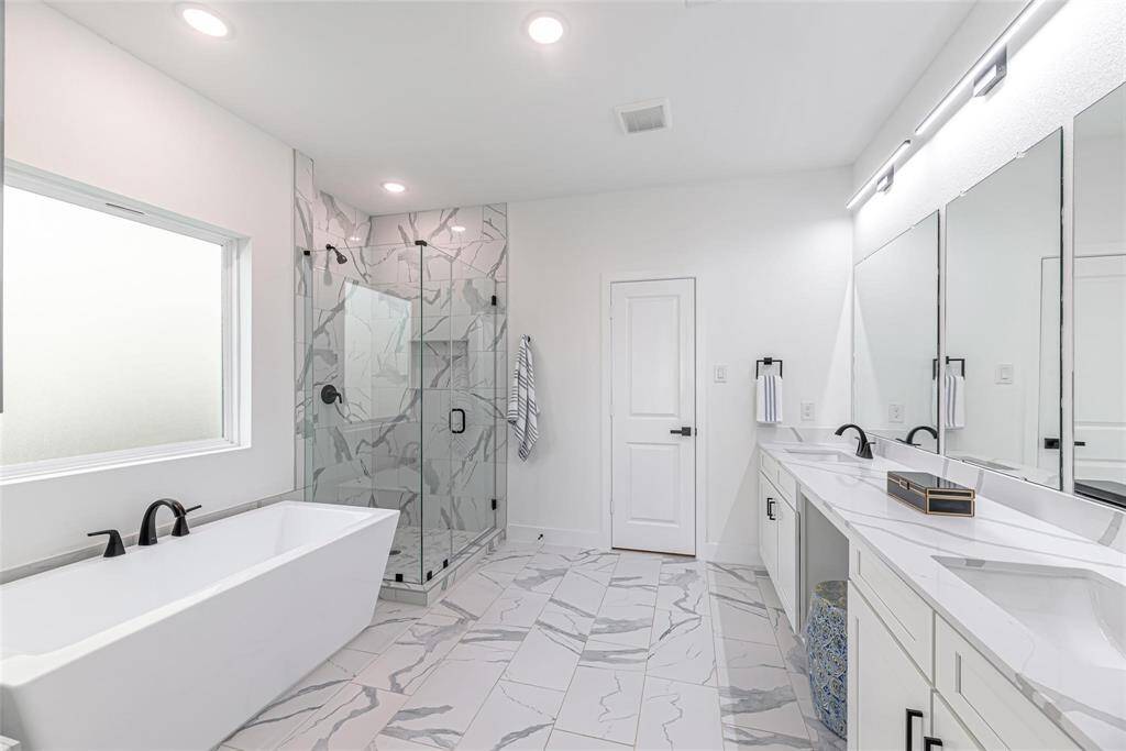 This primary bathroom features a modern design with dual sinks, sleek black fixtures, and ample white cabinetry. The large mirror enhances the space, and the glass-enclosed shower boasts stylish marble-patterned tiles. Bright lighting and a clean aesthetic complete the look.
