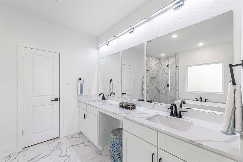This primary bathroom features a modern design with dual sinks, sleek black fixtures, and ample white cabinetry. The large mirror enhances the space, and the glass-enclosed shower boasts stylish marble-patterned tiles. Bright lighting and a clean aesthetic complete the look.