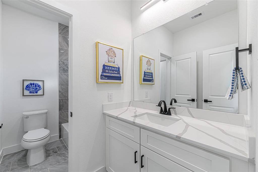 This modern bathroom features a sleek vanity with a large mirror, black fixtures, and ample storage. The room includes a toilet and a shower area with elegant tile work. Bright and stylish decor adds a touch of luxury.