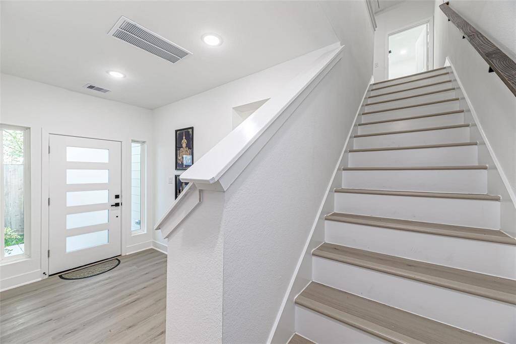 A light-filled, contemporary foyer with a staircase that leads to the upper level. The room has a modern and welcoming air thanks to its white walls, light wood floors, and a sleek entrance door with frosted glass panels.