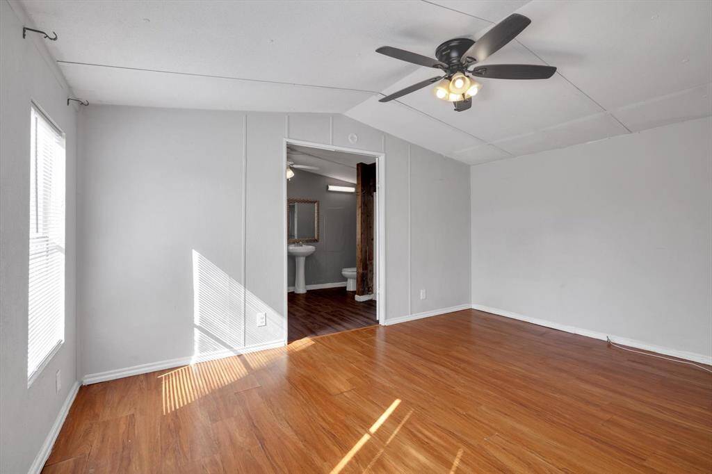 This room features sleek flooring, neutral gray walls, and a ceiling fan with lights. It has a doorway leading to a bathroom with a pedestal sink, offering a spacious and modern feel. Natural light streams in through the window, enhancing the bright and airy atmosphere.