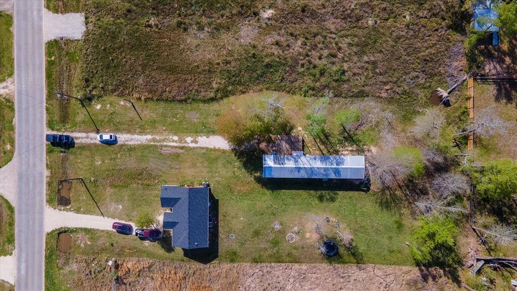 Property with a single-story house and a long outbuilding, set on a spacious lot with grassy areas and a few trees. The property is bordered by a road and features a driveway with parked vehicles. It's a rural setting with ample outdoor space.
