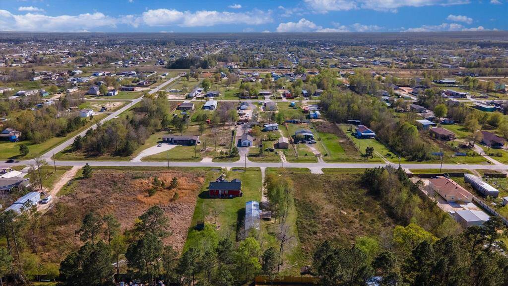 Spacious suburban area with scattered homes, open lots, and tree-lined streets. The neighborhood appears peaceful and expansive, offering a blend of greenery and residential development under a clear blue sky.