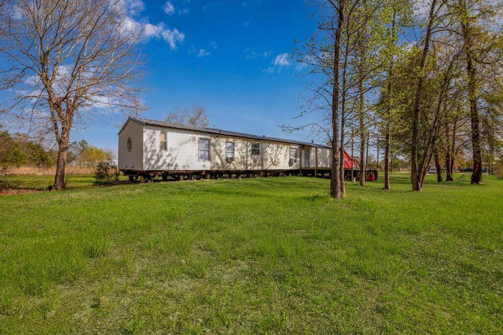 Mobile home elevated on a green, spacious lot with several trees, under a clear blue sky, offering potential for outdoor enjoyment and privacy.