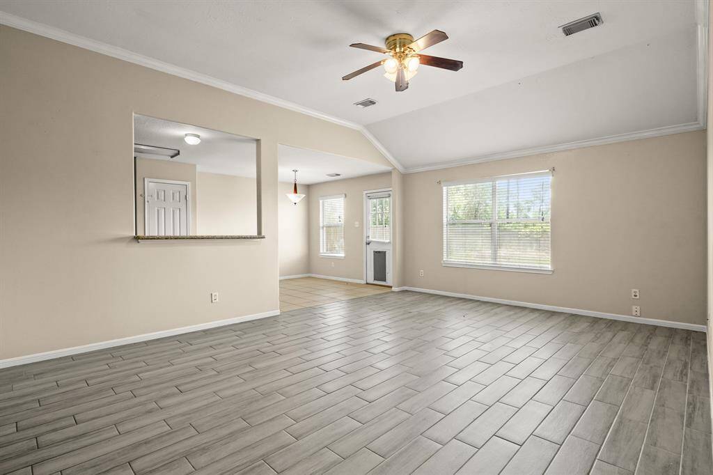 Spacious living area with wood like flooring and ample natural light from a large window that views to the backyard. Features a ceiling fan and a pass-through to the kitchen, enhancing the open feel.