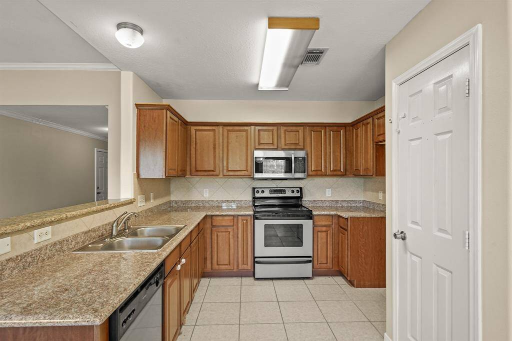 This kitchen features granite countertops, stainless steel appliances, and warm wood cabinetry. It has a double sink, a built-in microwave, and an electric stove, with a tile floor and an open layout connecting to the main living area.