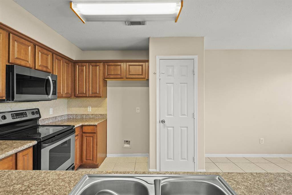 This kitchen features warm wooden cabinets, granite countertops, and stainless steel appliances. It has a built-in microwave, electric stove, and a double sink with a view of a spacious pantry area.