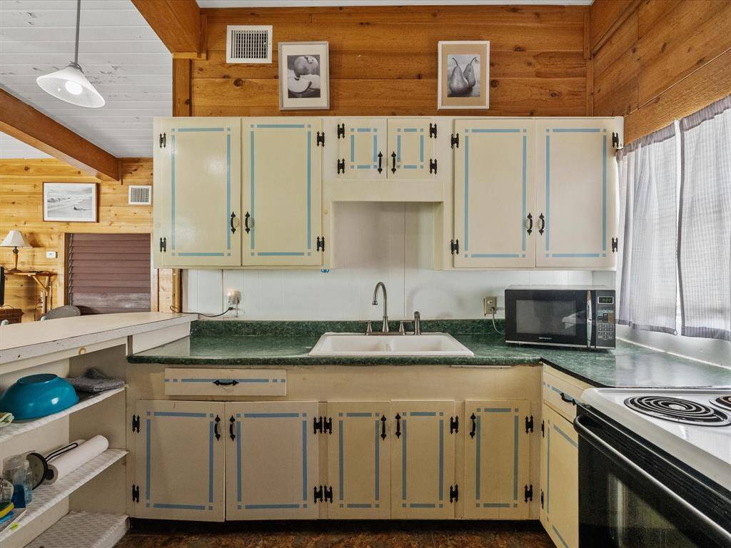 Cozy kitchen with rustic wood paneling and vintage charm. Features include painted cabinets, a green countertop, and essential appliances. Open shelving and a window provide natural light.