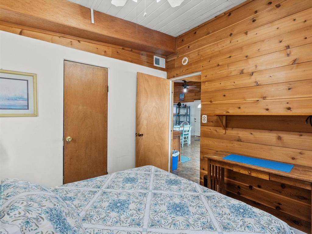 Cozy bedroom with wood paneling, featuring a built-in shelf and desk. It has a door leading to an adjoining room with a view of a dining area. The space is adorned with a floral-patterned bedspread and a framed picture on the wall.