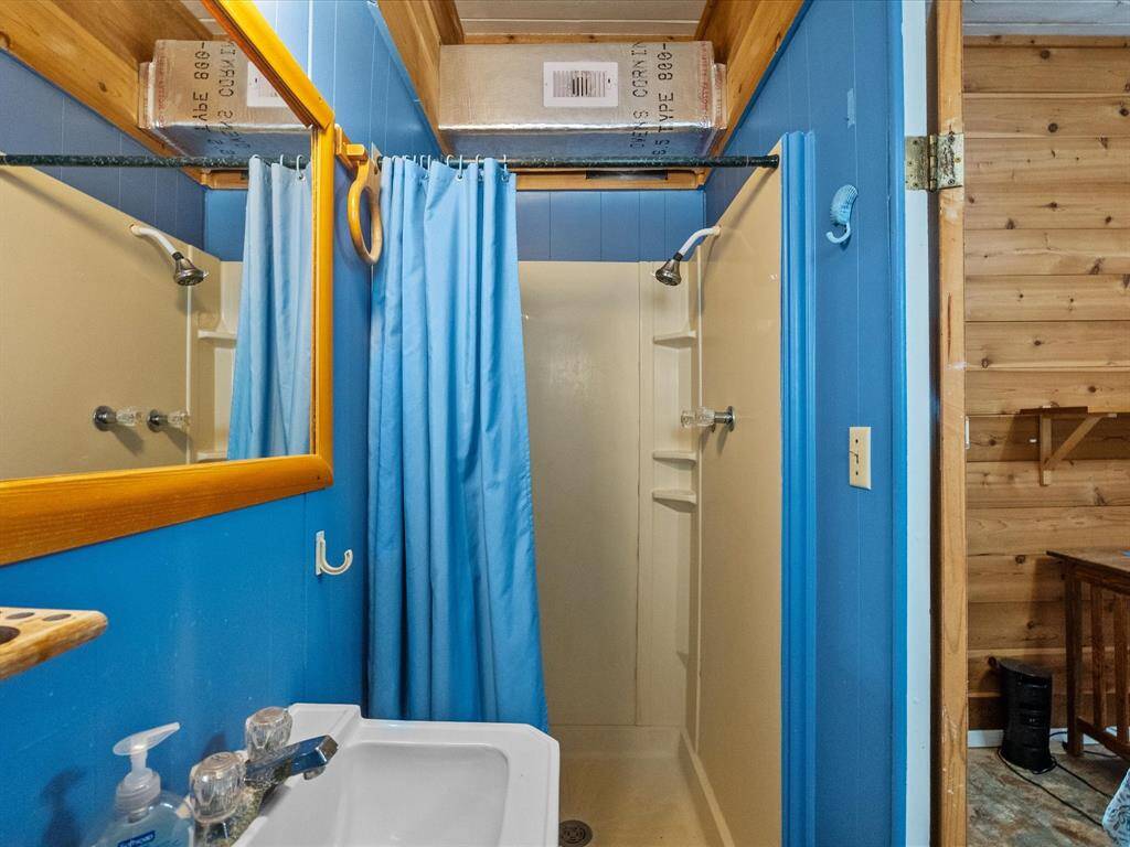 This bathroom features a compact shower with a blue curtain, matching blue walls, a small sink with a wooden-framed mirror, and wood paneling visible in the adjacent room.