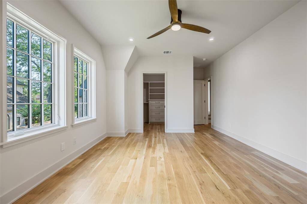 This sunlit secondary bedroom blends comfort with luxury, featuring rich hardwood flooring, recessed lighting, and a modern ceiling fan. The custom walk-in closet offers generous storage, while large windows provide treetop views and a peaceful ambiance throughout the day.
