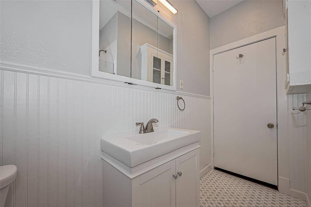 This photo shows a compact laundry area with a stacked washer and dryer, located in a hallway with light gray walls and dark wood flooring. It features a wall-mounted light fixture and two closed doors adjacent to the laundry space.
