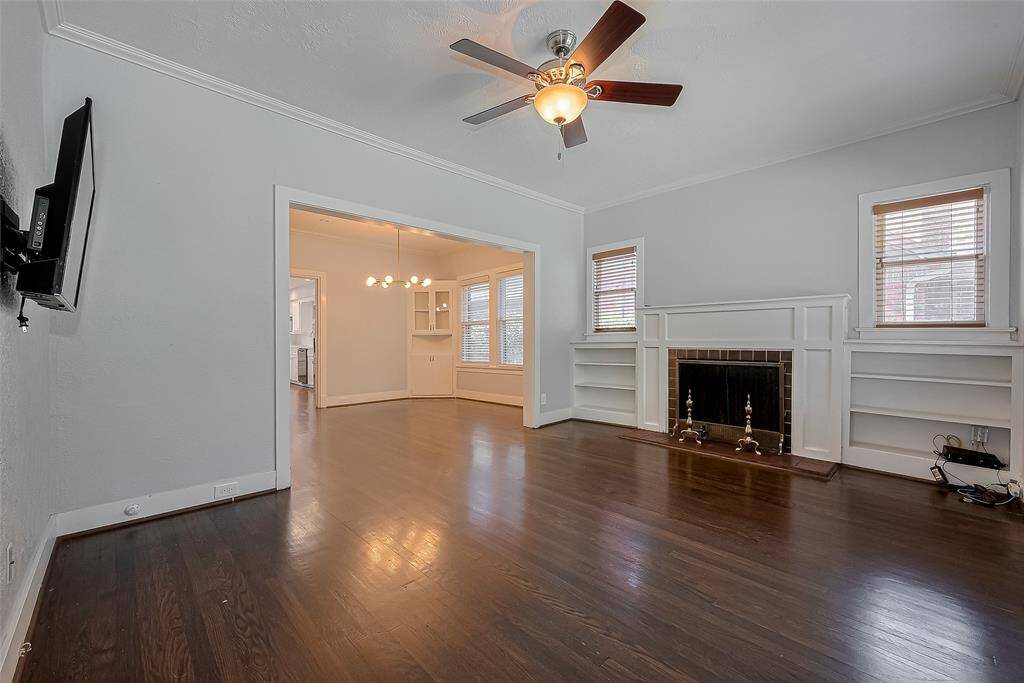 This living room features hardwood floors, a ceiling fan, built-in shelving, and a cozy fireplace. Large windows provide natural light, and a unique round-top door adds charm.