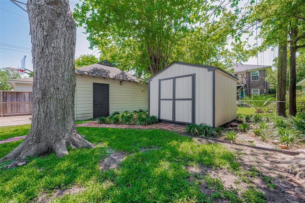 This photo showcases a spacious backyard with a covered wooden pergola, perfect for outdoor dining with a picnic table beneath. The yard includes a grassy area, a garage, a basketball hoop, and a storage shed, all enclosed by a wooden fence with greenery.