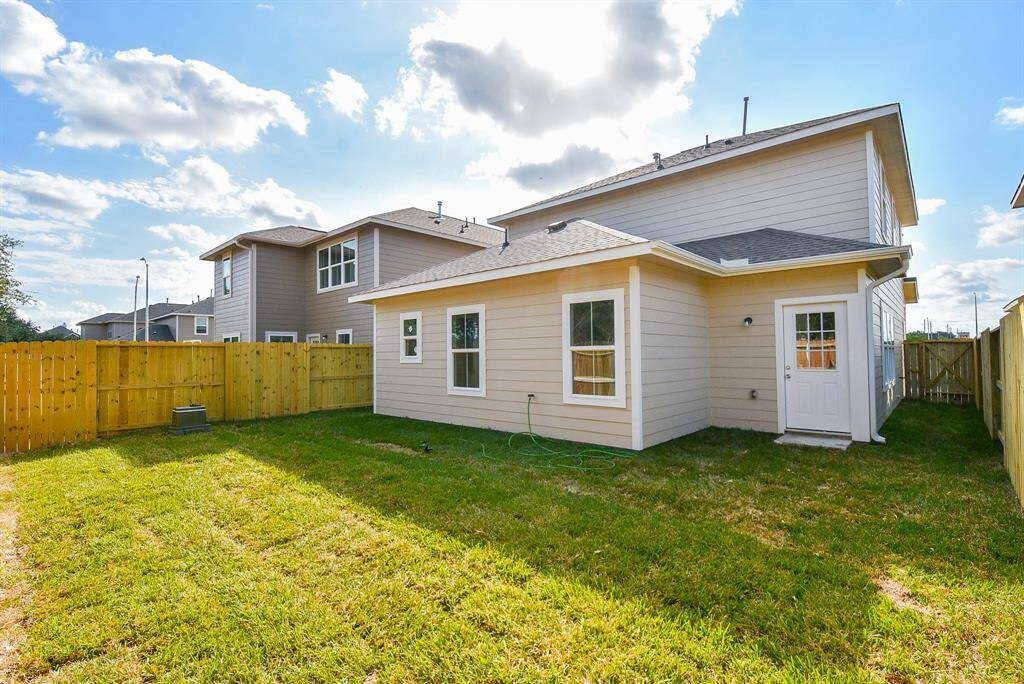 The photo shows a spacious backyard with a well-maintained lawn and a wooden privacy fence. The back of a two-story house with a modern design, featuring a mix of siding materials and a small back door, is visible. The yard offers ample space for outdoor activities and gardening.