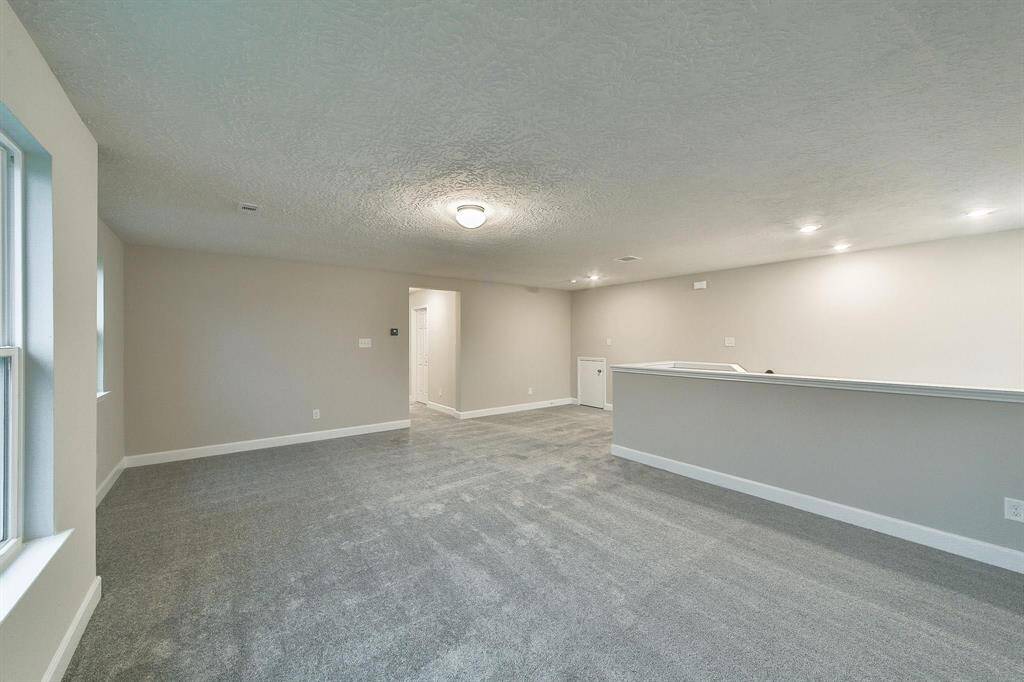 This is a spacious, well-lit room with neutral walls and gray carpet flooring, featuring multiple recessed lights and a window for natural light. The room includes a half-wall ledge that could serve as a division or a bar area.