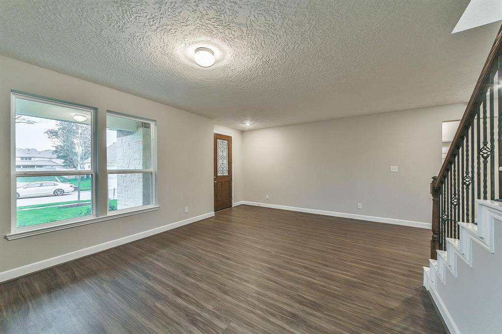 This is a bright, spacious room featuring large windows, wood-look flooring, a textured ceiling, and a staircase with a classic railing design.