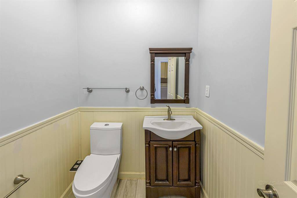 This photo shows a compact bathroom with light walls and wainscoting. It features a modern toilet, a dark wood vanity with a sink, and a matching framed mirror. There's also a towel holder on the wall.