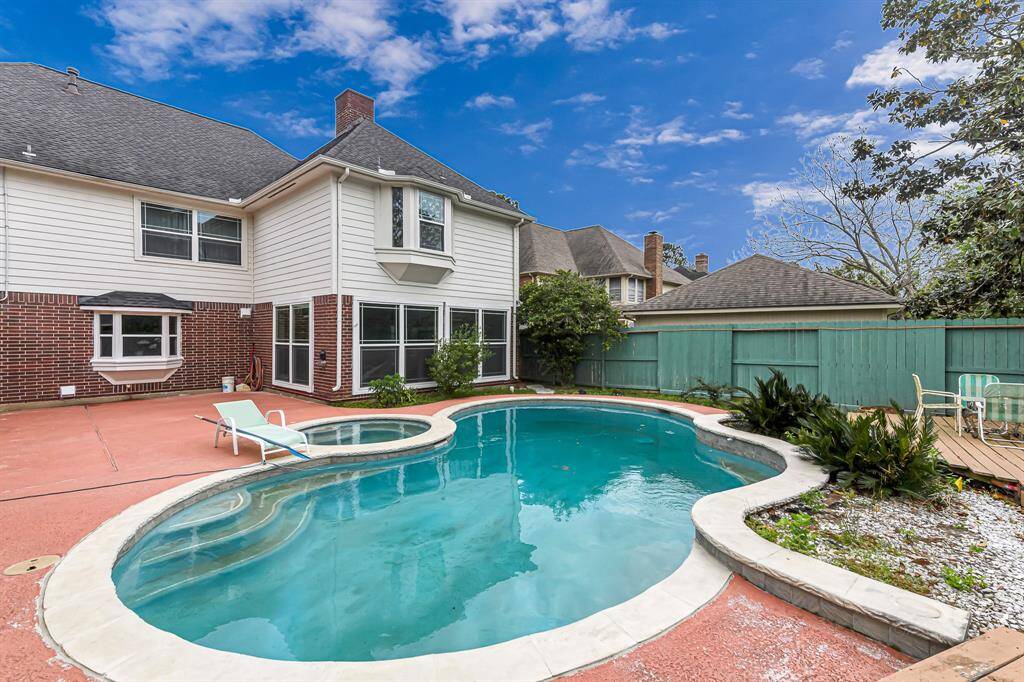 This photo showcases a spacious backyard with a uniquely shaped swimming pool and a hot tub. The area features a red concrete patio, a privacy fence, and some greenery. The house exterior is brick and siding, offering large windows that provide a view of the pool area, perfect for entertaining or relaxing.