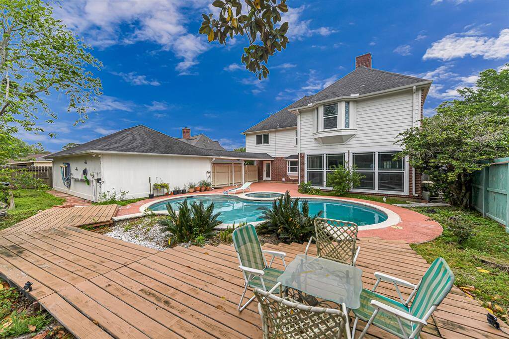 Spacious backyard with a large two-story house featuring a bay window. There's a circular pool surrounded by a red brick patio, a wooden deck with seating, and lush greenery, perfect for outdoor relaxation.