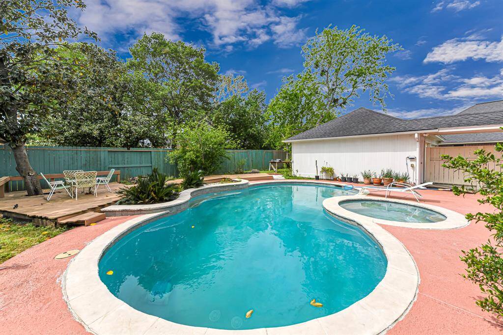 This backyard features a spacious pool with a connected spa, set against a backdrop of lush greenery and a privacy fence. There's a cozy deck area with seating, perfect for outdoor relaxation and entertaining.