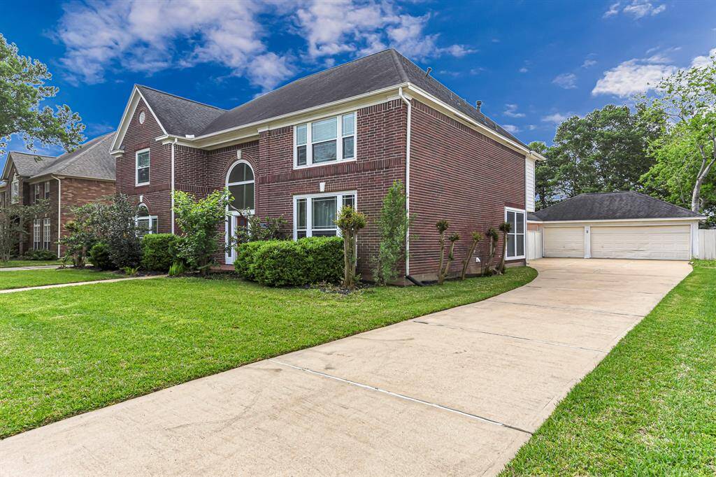 This photo shows a spacious two-story brick home with a well-maintained lawn and a long driveway leading to a detached two-car garage. The property is surrounded by greenery, offering a welcoming and serene atmosphere.