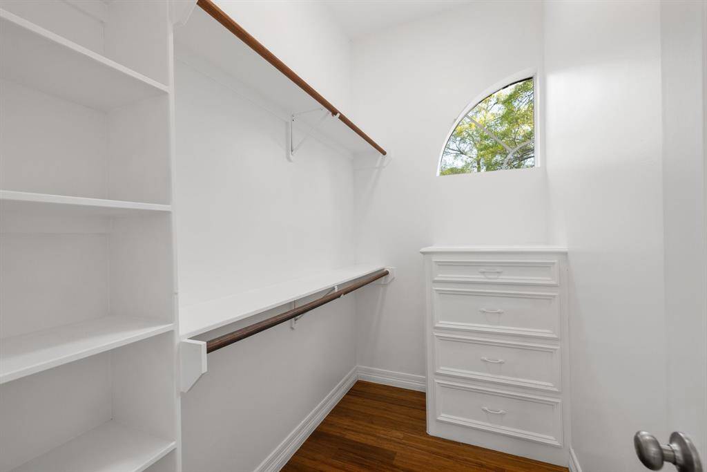 This photo shows a bright, well-organized walk-in closet with built-in shelving and a dresser. It features a wooden rod for hanging clothes and a small arched window that brings in natural light. The space has white walls and a wooden floor.