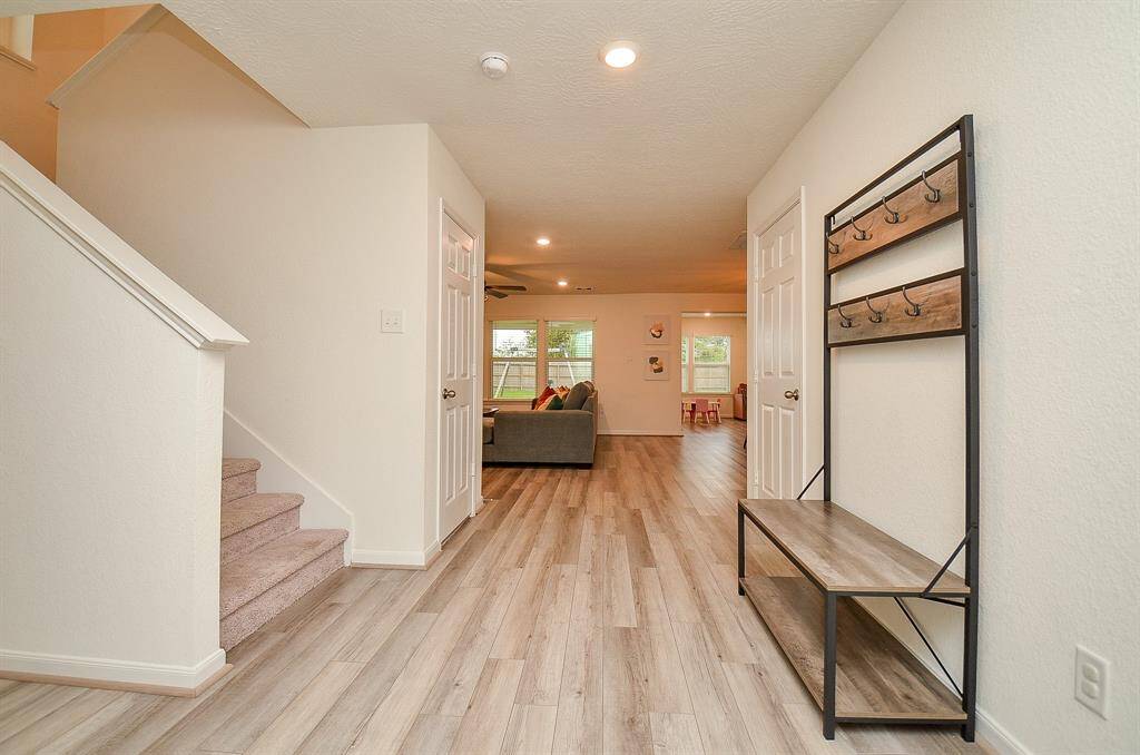 This photo showcases a welcoming entryway with light wood lam. flooring and neutral walls. A staircase leads upstairs, while the hallway opens into a cozy living space with ample natural light.