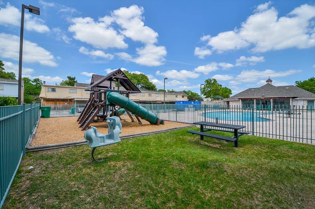 This photo showcases a community area with a fenced playground, featuring a slide and climbing structure, alongside a swimming pool and picnic bench, ideal for family recreation.