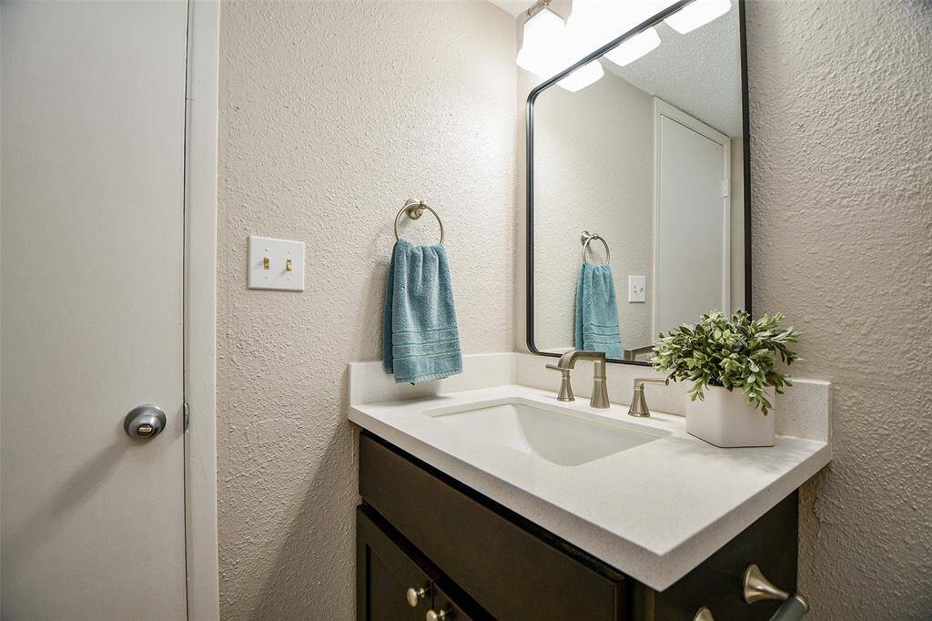 This bathroom features a modern vanity with a sleek countertop and stylish faucet. It includes a large mirror, overhead lighting, and a decorative plant. A towel ring with a blue towel adds a pop of color.