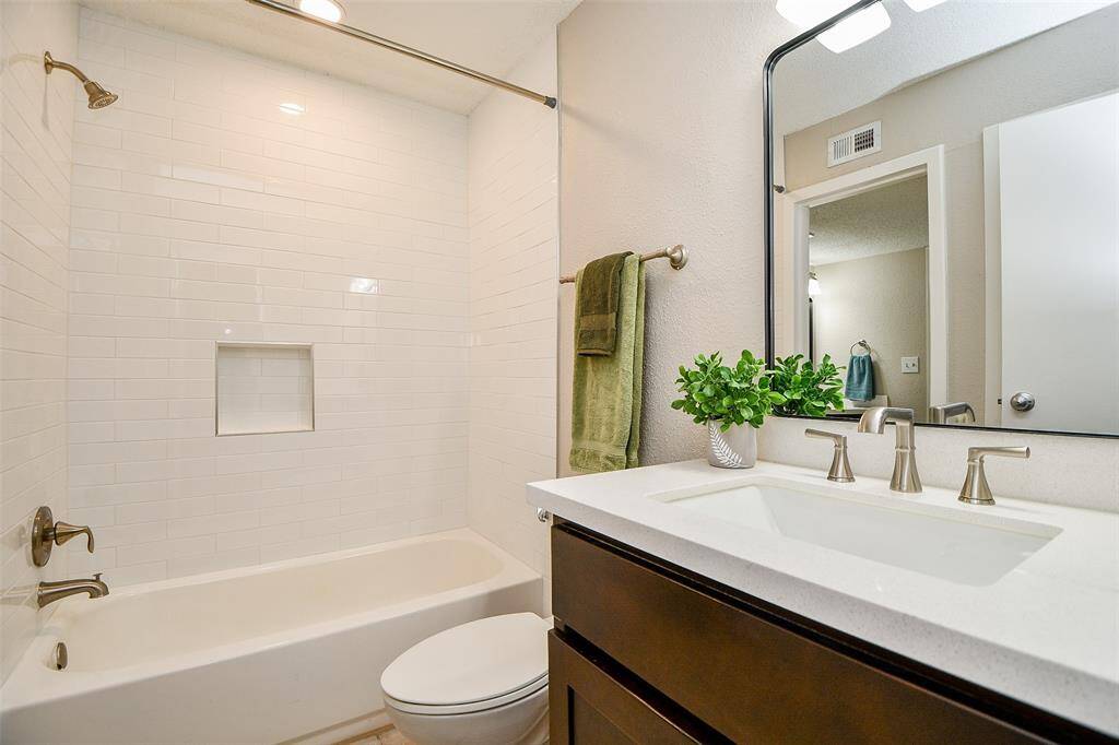 This modern bathroom features a sleek vanity with a large mirror, a spacious white bathtub with subway tile surround, and elegant fixtures. It includes a built-in shelf and is accented by a touch of greenery for a fresh feel.