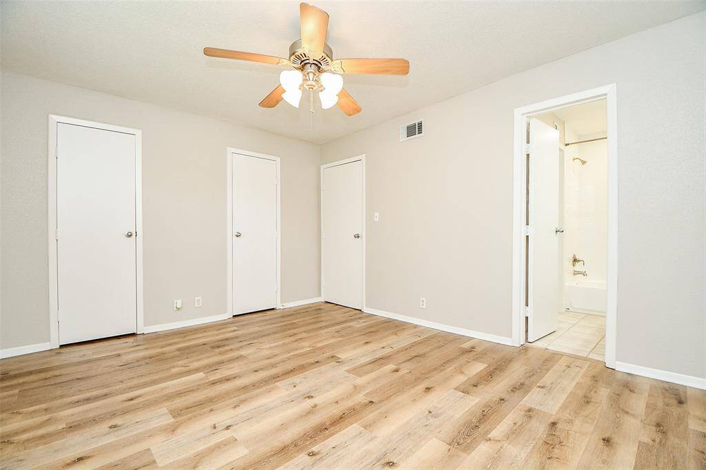 Bright bedroom with light wood flooring, neutral walls, ceiling fan, and three white doors. One door leads to an en-suite bathroom with a tub.