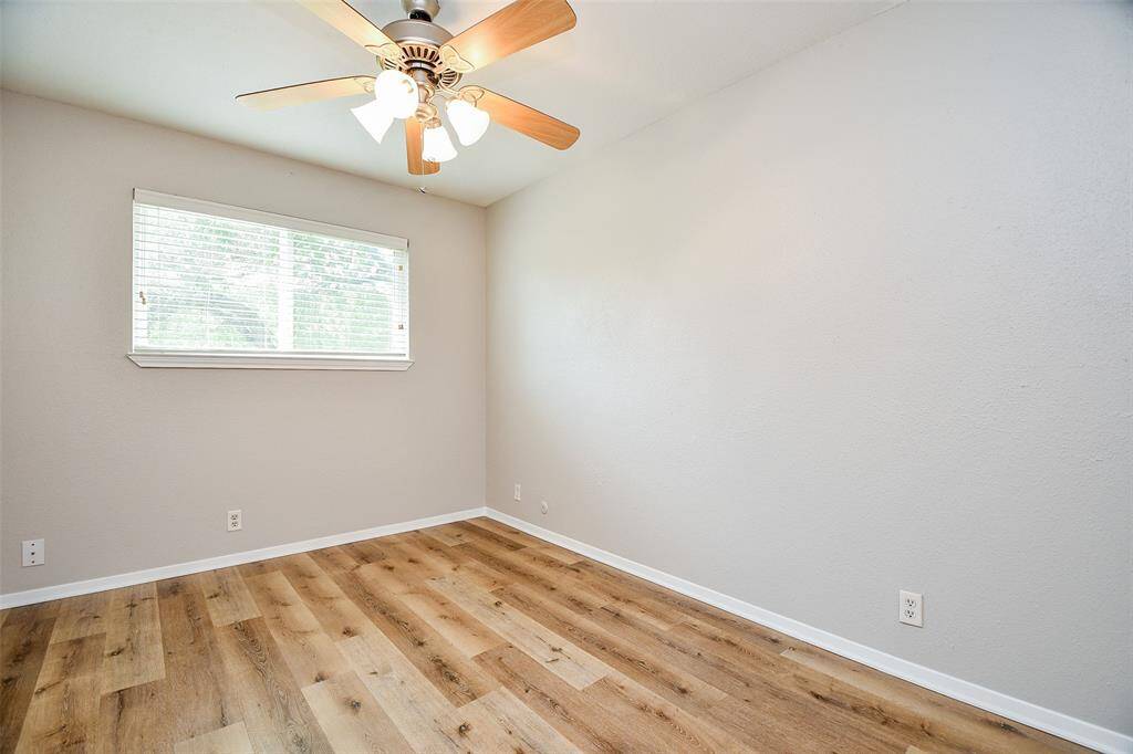 This bright room features fresh light gray walls, wood-style flooring, a large window with blinds, and a ceiling fan with lights, creating a welcoming and airy atmosphere.