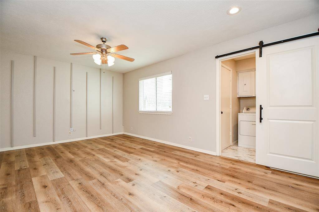 This room features laminate wood flooring, a ceiling fan, and a modern barn door leading to a laundry area. The neutral walls with vertical accents add a touch of style, and a large window provides natural light.