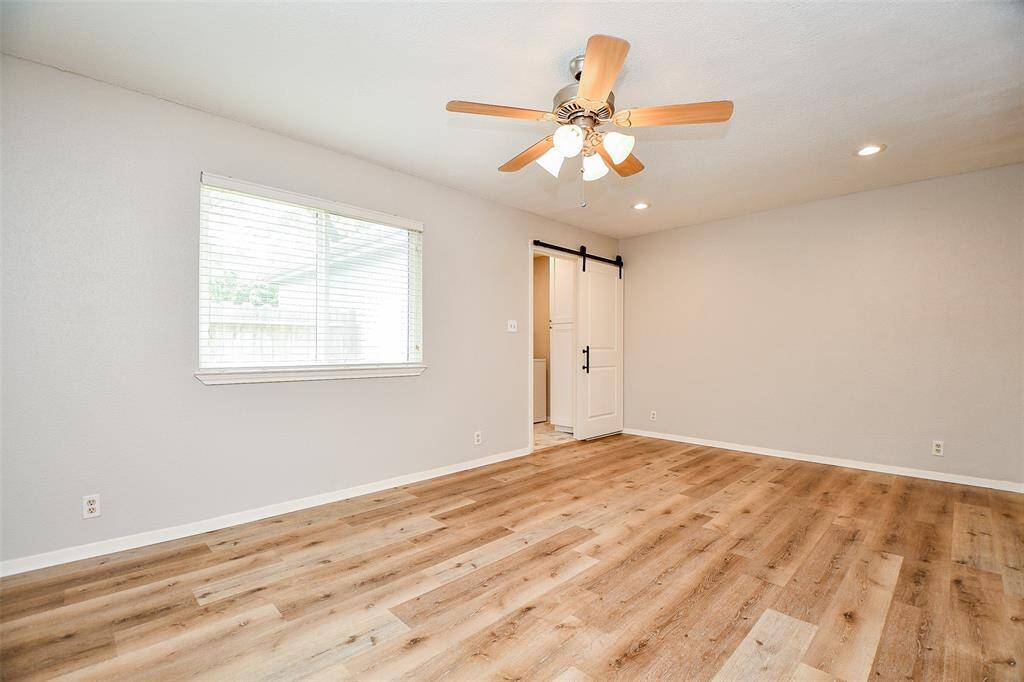 This room features a bright, airy space with light wood flooring and neutral walls. It includes a ceiling fan, recessed lighting, and a large window. A sliding barn door adds character and leads to another area.