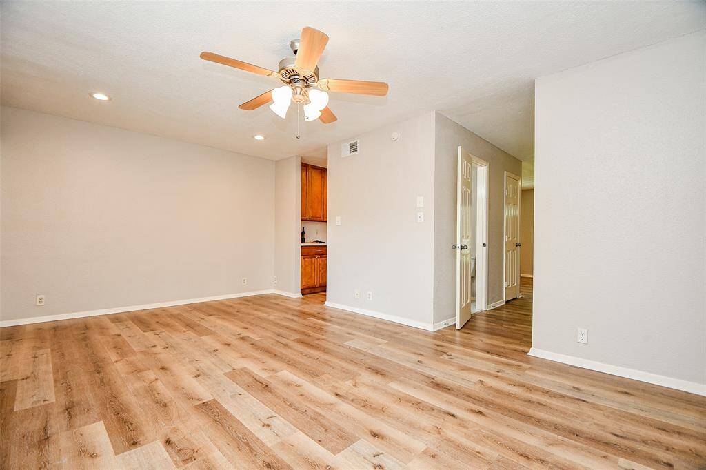 This spacious living area features modern wood-look flooring, neutral walls, and a ceiling fan. The open layout leads to a partially visible kitchen, creating a welcoming and functional space.
