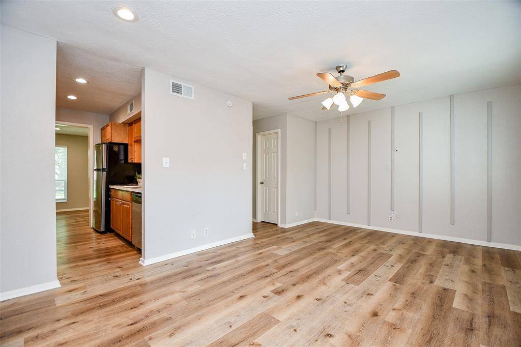 This room features modern wood flooring, neutral walls with vertical accents, and a ceiling fan with lights. The open layout connects to a kitchen with wooden cabinets and stainless steel appliances, enhancing the bright, spacious feel.