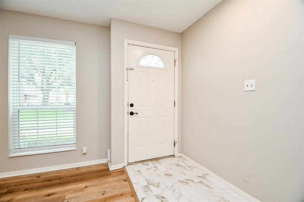 Bright entryway with a mix of wood and tile flooring. The space features a large window with blinds, offering natural light, and a white front door with decorative glass detail. Neutral wall color enhances the welcoming feel.
