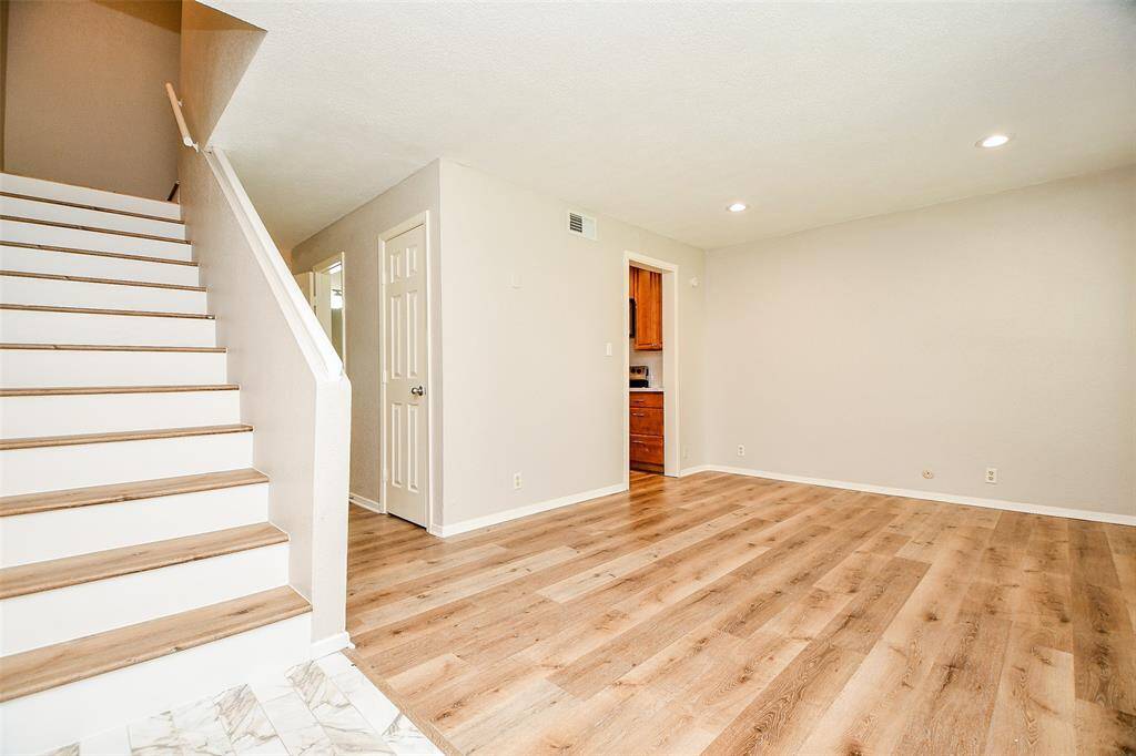 This inviting space features modern wood flooring, neutral walls, and recessed lighting. A staircase with wooden steps leads to the upper level, and a doorway opens to a cozy kitchen area, providing a warm and open feel.