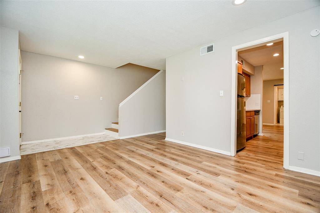 This space features an open living area with modern wood flooring, neutral walls, and recessed lighting. A staircase leads to the upper level, and there's a view into a kitchen with stainless steel appliances.