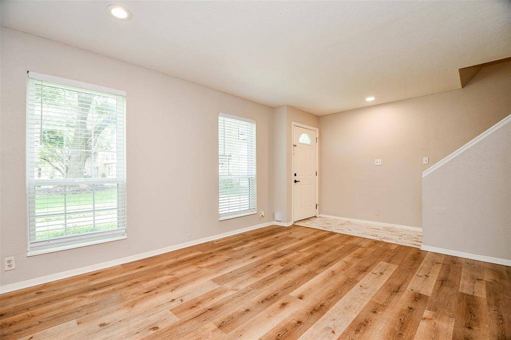 Bright and inviting living area with hardwood floors, neutral walls, and large windows. The space features recessed lighting and an entryway with stairs, providing a welcoming atmosphere.
