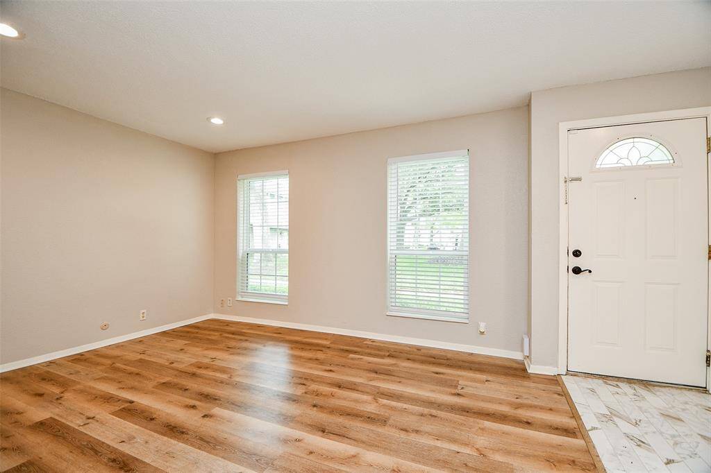 This room features a bright, open space with two windows, hardwood flooring, and a neutral color palette. The entryway has a tiled area, and the room is well-lit with recessed lighting.