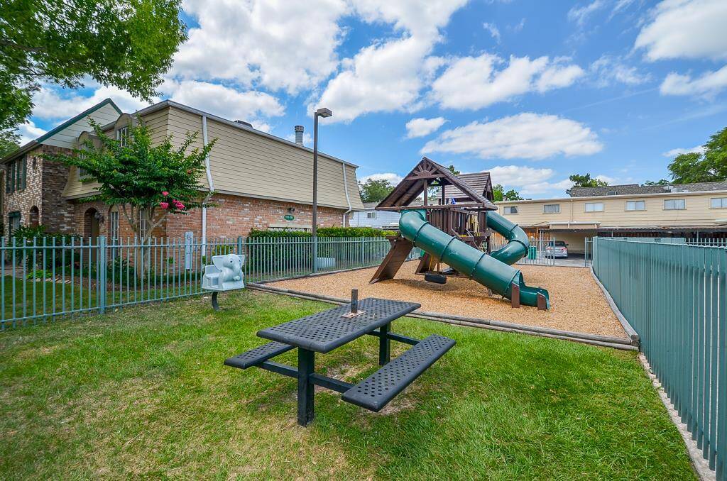 This photo showcases a quaint backyard with a fenced playground area, featuring a slide and climbing structure. There's a picnic table on the grassy section, perfect for outdoor gatherings. The surrounding buildings suggest a family-friendly community setting.