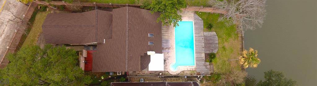 This aerial view shows a spacious property featuring a large pool and deck area, adjacent to a body of water. The house is surrounded by mature trees, providing privacy and a natural setting.
